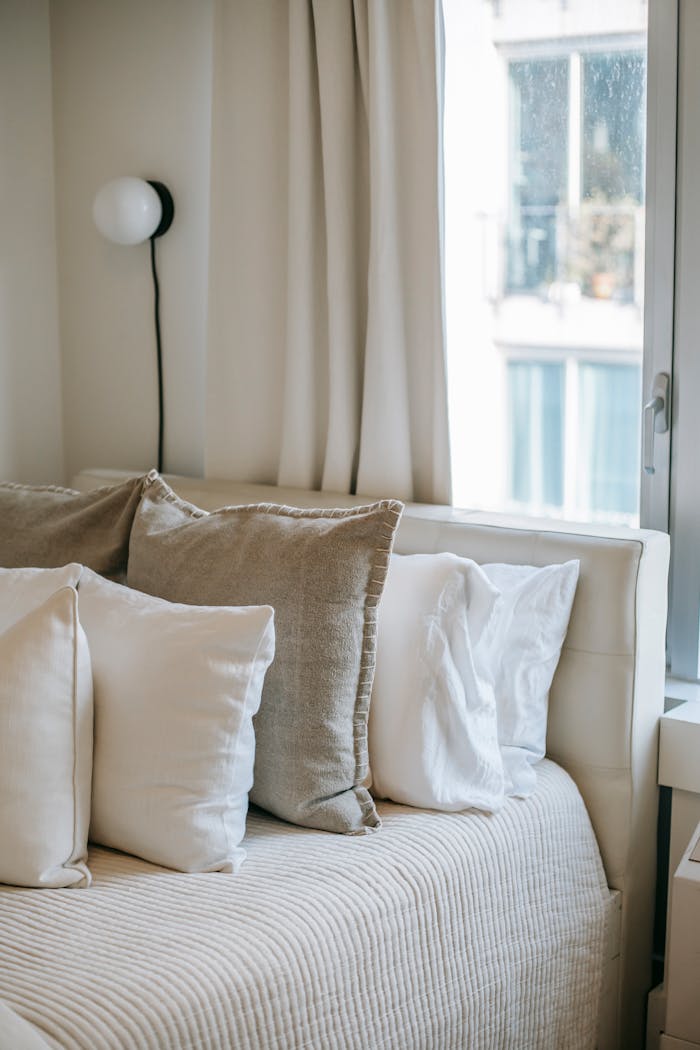 Cozy bed with assorted pillows on cover near shiny window and lamp on wall in house