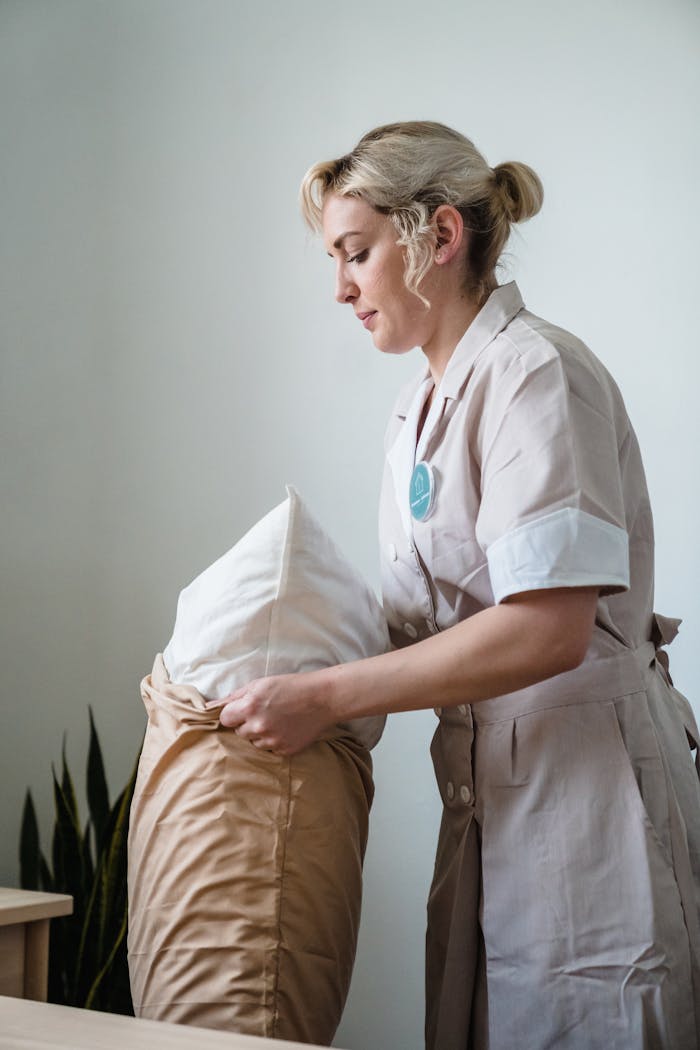 services-05 Hotel maid in uniform arranging pillows in a neatly styled guest room.